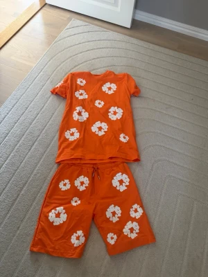 Orange blommigt set med t-shirt & shorts - Säljer ett orange set med t-shirt och shorts, dekorerat med stora vita blommor. Perfekt för dig som gillar färgstarka och lekfulla plagg. Både tröjan och shortsen har avslappnad passform och är tillverkade i mjuk bomull.