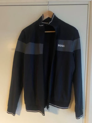 Mörkblå ziptröja från BOSS - Snygg mörkblå ziptröja från BOSS med gråa ränder över bröstet och armarna. Tröjan har hög krage, dragkedja framtill och vita detaljer vid krage, ärmslut och nederkant. Perfekt för en stilren och sportig look.