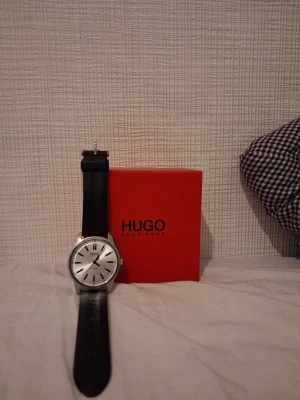 Hugo Boss silverfärgad klocka - Stilren klocka från Hugo Boss med silverfärgad urtavla och svart armband. Klockan har minimalistisk design med tydliga svarta markeringar och klassisk rund form. Levereras med originalförpackning i röd box.                              -Pris går att diskuteras