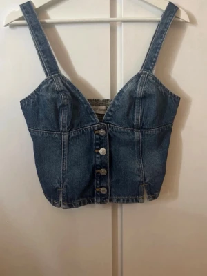Jeans korsettopp med knappar - Trendig jeans korsettopp med breda axelband och knappar framtill. Toppen har en croppad passform och smockad rygg för extra komfort. Perfekt för dig som vill ha en edgy och cool look till sommaren.