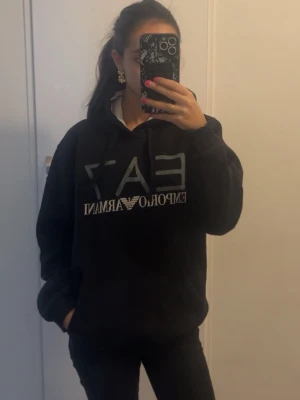 Svart EA7 Emporio Armani hoodie - Snygg svart hoodie från EA7 Emporio Armani med stor logga framtill. Hoodien har huva med dragsko, magficka och mjukt innerfoder i huvan som är vitt. Perfekt för en chill och trendig look.