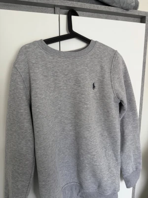 Grå sweatshirt från Ralph Lauren - Klassisk grå sweatshirt från Ralph Lauren med rund halsringning och diskret broderad logga på bröstet. Tröjan har ribbade muddar vid ärmslut och nederkant, perfekt för en clean och avslappnad stil. Tillverkad i mjukt material för extra komfort.