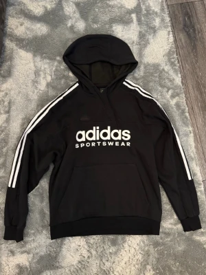 Svart Adidas hoodie Storlek S - Snygg svart hoodie från Adidas med klassiska vita ränder längs ärmarna och stor Adidas Sportswear-logga på bröstet. Huva och magficka framtill. Perfekt för en sportig och avslappnad stil.Storlek S