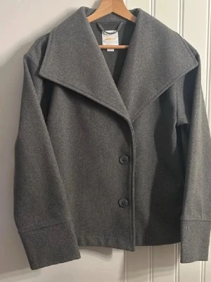 Grå kappa med bred krage - Stilren grå rock med bred krage och två stora knappar framtill. Tillverkad i mjuk ullmix som ger en klassisk look. Rak passform och långa ärmar, perfekt för kyliga dagar. Enkel men trendig design som passar till många outfits. Skicka ⭐ i meddelande för en hemlig rabatt.