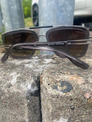  fyrkantiga solglasögon - Snygga solglasögon från Marc Jacobs med fyrkantiga bågar i mörkgrått och svarta detaljer. Glasen är tonade och ramen har en cool metallkänsla. Perfekt accessoar för att lyfta din stil under soliga dagar.
