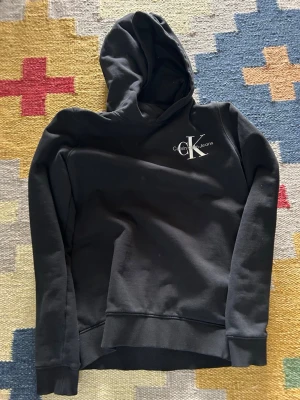 Svart hoodie från Calvin Klein Jeans - Svart hoodie från Calvin Klein Jeans med vit logga på bröstet. Klassisk modell med huva, långa ärmar och ribbade muddar. Tillverkad i mjukt material som känns skönt mot huden. Perfekt för en avslappnad och stilren look.