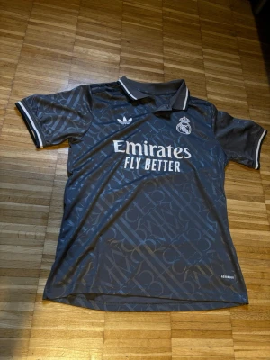 Real Madrid  svart matchtröja - Snygg svart Real Madrid matchtröja med vita detaljer och trycket 'Emirates Fly Better' på bröstet. Tröjan har ett diskret mönster, klubbmärke  AEROREADY-material för bra ventilation. Perfekt för fotboll eller träning.