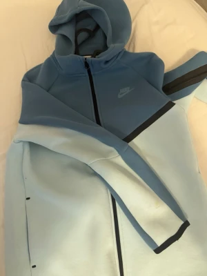 Nike Tech Fleece blå hoodie - Snygg Nike Tech Fleece hoodie i två nyanser av blått med hel dragkedja och svarta detaljer. Hoodien har huva, fickor med dragkedja och Nike-logga på bröstet. Tillverkad i mjukt och bekvämt fleecematerial, perfekt för en sportig och avslappnad stil.