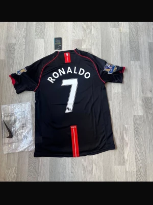 Manchester United Ronaldo tröja svart - Snygg svart Manchester United matchtröja från Nike med Ronaldo och nummer 7 på ryggen. Tröjan har röda detaljer, Premier League-patchar på ärmarna och AIG-logga på bröstet. Tillverkad i lätt och ventilerande material, perfekt för fotboll eller streetwear.