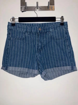 Randiga denimshorts från &Denim - Säljer ett par blå denimshorts från &Denim med vita vertikala ränder och regular waist. Shortsen har klassisk femficksdesign, knappgylf och uppvikta benslut. Perfekta för sommardagar och enkel att matcha med olika toppar.