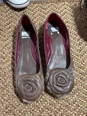 Bruna ballerinaskor med rosdetalj (37) - Unika bruna ballerinaskor  med stor rosdetalj framtill och fluffig kant. Insidan är vinröd och skon har rundad tå. Perfekta för dig som gillar detaljer och vill sticka ut lite extra.