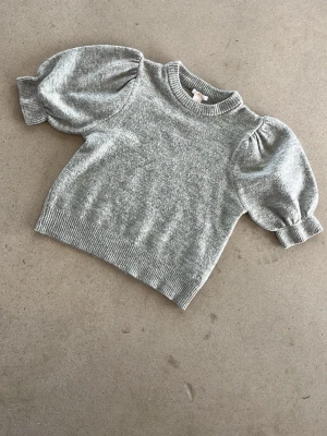 Grå stickad tröja med puffärm Gina Tricot - Grå stickad tröja från Gina Tricot med trendiga puffärmar och ribbade muddar vid hals, ärmslut och nederkant. Perfekt för dig som gillar en stilren look med en twist. Mjuk och skön kvalitet.
