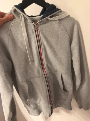 Ljusgrå hoodie från Pier One - Ljusgrå hoodie från Pier One med dragkedja och två fickor framtill. Huvan har vita dragsnören och dragkedjan har en snygg detalj med röd och marinblå rand. Perfekt för en avslappnad stil och enkel att matcha med jeans eller joggers.