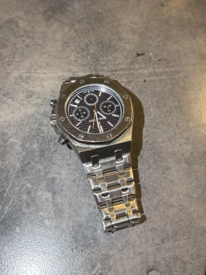 AP klocka silver - Snygg Audemars Piguet Royal Oak klocka i silverfärgat stål med ikonisk åttakantig boett och svart urtavla med tre subdials. Länkarmband i borstat stål och detaljerade skruvar runt boetten ger en lyxig och sportig känsla.