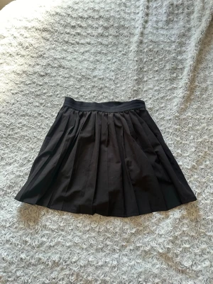 Svart plisserad skort - Snygg svart skort med plisserad kjoldel och inbyggda shorts. Skorten har en  midjeresår och är tillverkad i ett mjukt, stretchigt material. Skjortsen under har ett fack för en tennisboll samt en praktiskt ficka för mobilen. 