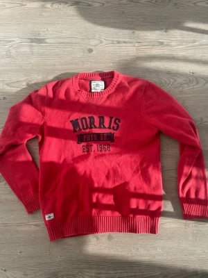 Morris tröja  - Röd stickad tröja från Morris med broderat tryck 'Morris Phys. Ed. Est. 1968' på bröstet. Tröjan har rund halsringning, ribbade muddar och en klassisk passform. Perfekt för dig som gillar sportig och preppy stil.