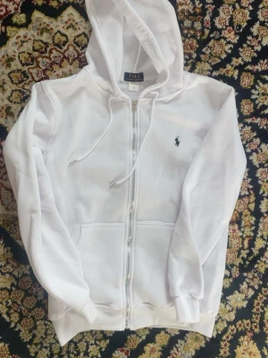 Vit hoodie från Polo Ralph Lauren - Snygg vit hoodie med dragkedja från Polo Ralph Lauren. Klassisk design med huva och dragsnören, två fickor framtill och den ikoniska lilla loggan broderad på bröstet. Perfekt för en clean och stilren look.
