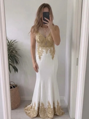 Fest klänning  - Elegant vit långklänning med detaljerad guldspets upptill och längs fållen. Klänningen har ärmlös design, mesh upptill och figurnära passform som ger en lyxig känsla. Perfekt för bal eller festliga tillfällen.