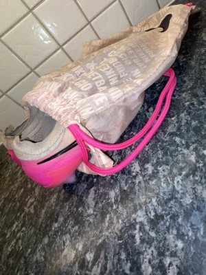 Nike rosa och gråa fotbollsskor - Säljer ett par snygga fotbollsskor från Nike i grått och rosa med svarta detaljer. Skorna har en modern design med dobbar för bra grepp på planen och kommer med en matchande Nike-påse. Perfekta för dig som vill sticka ut på fotbollsplanen.