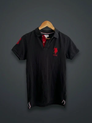 US Polo Assn. Pikétröja – Svart med röda detaljer - Stilren och klassisk pikétröja från US Polo Assn. i svart färg med röda detaljer och broderad logga på bröstet. Perfekt till vardag, sommar, semester eller en avslappnad men välklädd look. Matchas enkelt med jeans, chinos eller shorts.  ✅ US Polo Assn. original ✅ Svart med röda detaljer ✅ Broderad logga framtill ✅ Bekväm passform ✅ Klassisk pikékrage ✅ Fint använt skick  📏 Storlek: S 📍 Skick: mycket bra   Skicka gärna meddelande vid frågor eller intresse!