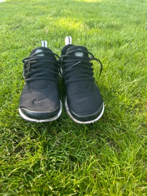 Svarta Nike Presto sneakers - Säljer ett par svarta Nike Presto sneakers med vit sula och diskret Nike-logga på tån. Skorna har en låg profil, mesh-material och plastdetaljer på sidan för extra stöd. Perfekta för dig som gillar sportig och clean stil.
