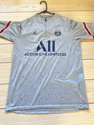 PSG grå matchtröja Jordan x Nike - Snygg grå PSG matchtröja med svarta prickar, blå logga och texten 'Accor Live Limitless' på bröstet. Röd, vit och svart rand på ärmarna, PSG-märke och Jordan-logga framtill. Tillverkad i lätt och ventilerande funktionsmaterial. Har använts 2 gånger