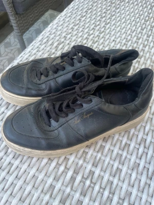 Svarta Axel Arigato sneakers i läder - Snygga svarta sneakers från Axel Arigato i äkta läder med svart snörning och diskret logga i guld på sidan. Skorna har en klassisk låg siluett och vit sula som ger en clean look. Perfekta för dig som gillar minimalistisk och stilren design.