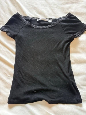 Svart topp med spetsdetaljer - Snygg svart topp med korta ärmar och fina spetsdetaljer vid halsringning och ärmslut. Modellen är figurnära och passar perfekt till både jeans och kjol. Enkel men ändå elegant design som ger en trendig look.