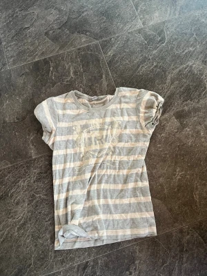 Randig kortärmad t-shirt i bomull - Säljer en ljusgrå och vit randig t-shirt med korta ärmar. T-shirten har ett diskret tryck framtill och är tillverkad i mjuk bomull. Perfekt för en avslappnad stil och enkel att matcha med jeans eller shorts. (Plus frakt)