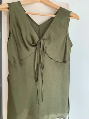 Olivgrön ärmlös topp med knyt - Snygg olivgrön ärmlös topp med v-ringning och knyt framtill. Klänningen har en lätt glansig yta och slits i sidan, vilket ger en modern och stilren look. Perfekt för varma dagar och enkel att styla med accessoarer.