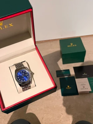 Role Datejust blå urtavla stål - Säljer en stilren Role Datejust med blå urtavla och silverfärgat stålarmband. Klockan har en räfflad bezel och index med stenar, samt datumvisning. Kommer i originalbox med tillhörande certifikat och manualer.