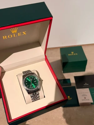Role klocka med grön urtavla - Exklusiv Role klocka med grön urtavla och silverfärgat armband i metall. Klockan har en bezel dekorerad med stenar och levereras i originalbox med certifikat och tillbehör. Perfekt för dig som vill ha något riktigt lyxigt på handleden.