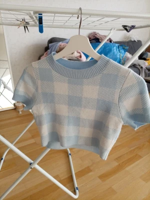 Blåvit rutig topp från Divided - Superfin blåvit rutig topp från Divided med korta ärmar och ribbad rund hals. Den har en croppad passform och är gjord i mjukt stickat material. Perfekt för dig som gillar en söt och trendig stil.