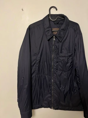 Oscar Jacobson Loden Nylon Jacket - Mörkblå lättviktsjacka från Oscar Jacobson i nylon. Stilren och minimalistisk modell med zip framtill och klassisk krage. Perfekt som vår-/sommarjacka eller för lager på lager  Aldrig använd   Märkt strl 50 (motsvarar L men sitter mer som en M enligt mig)