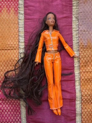 Vintage Barbie-docka med orange Y2K-outfit – tidigt 2000-tal - Vintage Barbie-docka från tidigt 2000-tal med lång mörkbrun hårsvall och iögonfallande orange outfit i äkta Y2K-stil. Dockan bär matchande jacka, byxor och skor med silverdetaljer som ger en klassisk tidig 2000-talslook.  Dockan är i fint vintage-skick med välbevarat ansikte, hår och kläder. Perfekt för samlare, nostalgiker eller den som gillar retro Barbie och Y2K-estetik.  Alla delar som syns på bilderna ingår.  Skick: Mycket bra