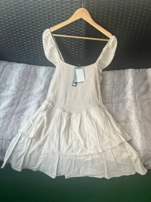 Beige volangklänning med puffärm - Supersöt beige kortklänning med volanger och puffärmar. Klänningen har smockad överdel och är tillverkad i ett lätt linnematerial, perfekt för varma dagar. Den har en romantisk och luftig känsla med fina detaljer och en feminin siluett.