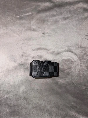 Svart rutigt LV-bälte - Snyggt svart bälte från Louis Vuitton med klassiskt rutigt mönster och stor LV-logga på metallspännet. Bältet har en exklusiv känsla och detaljer i svart metall. Perfekt accessoar för att lyfta din outfit.