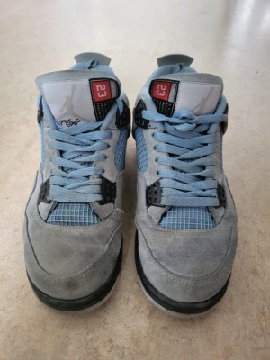 Air Jordan 4 University Blue sneakers - Snygga Air Jordan 4 University Blue sneakers med ljusblå mocka, meshpaneler och svarta detaljer. Skorna har klassisk Jordan 4-design med synlig Air-sula och coola plastdetaljer vid snörningen. Perfekta för dig som gillar streetwear och vill sticka ut.