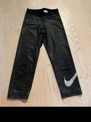 Svarta Nike leggings med logga - Snygga svarta leggings från Nike med stor vit Swoosh-logga på ena benet. Tillverkade i ett glansigt, stretchigt material som sitter tajt och bekvämt. Perfekta för träning eller chill. Midjan är elastisk för extra komfort.