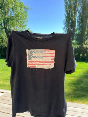 T-shirt denim Ralph Lauren - Svart t-shirt från Ralph Lauren Denim & Supply med en stor amerikansk flagga i sliten look framtill. Flaggan är sydd som en patch och har råa kanter för en cool vintagekänsla. Kom gärna med prisförslag och skriv vid minsta fundering! 💭😉😁😁