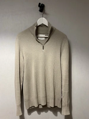 Beige stickad tröja med halv dragkedja - Snygg beige stickad tröja med ribbad struktur och halv dragkedja vid halsen. Tröjan har lång ärm och hög krage som kan vikas ner. Perfekt för lager på lager och enkel att matcha med olika outfits.