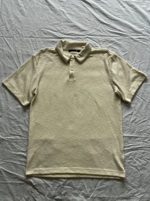 Beige polotröja  - Säljer en beige polotopp från Cubus med klassisk krage och två knappar framtill. Toppen är kortärmad och har en mjuk, strukturerad yta som ger en avslappnad men ändå stilren look. Perfekt att matcha med jeans eller kjol.