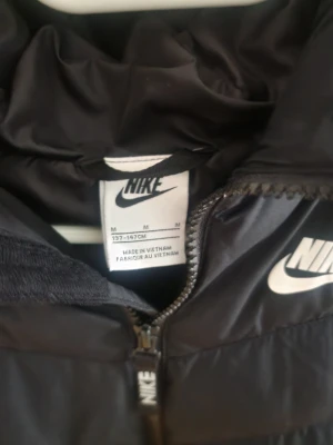 Svart dunjacka från Nike med huva - Snygg svart dunjacka från Nike med huva och dragkedja framtill. Jackan har två sidofickor och tydliga Nike-loggor på bröstet och dragkedjan. Perfekt för kalla vinterdagar och ger en sportig look.
