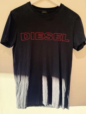 Svart t-shirt från Diesel - Säljer en svart t-shirt från Diesel med stort rött logotryck över bröstet. Klassisk rund halsringning och korta ärmar. Perfekt för dig som gillar enkel men snygg streetwear.