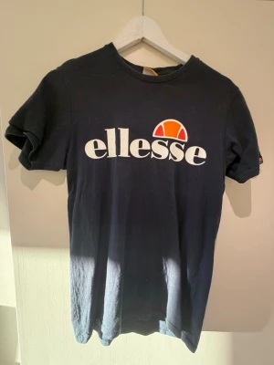 Svart t-shirt från Ellesse - Cool svart t-shirt från Ellesse med stort vitt logotryck och orange detalj på bröstet. Klassisk rund hals och korta ärmar, perfekt för en avslappnad och sportig look. Materialet är mjukt och bekvämt, troligtvis bomull.