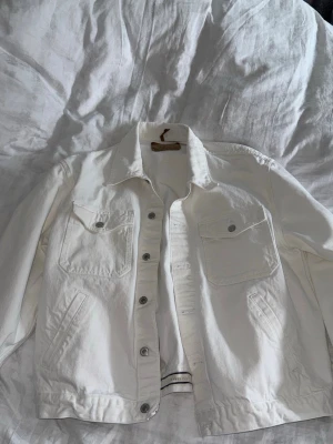 Vit jeansjacka från Jeanerica - Stilren vit jeansjacka med klassisk krage, två bröstfickor med lock och knappar samt sidofickor. Jackan har silverfärgade knappar framtill och raka ärmar. Perfekt för dig som vill ha en enkel men snygg jeansjacka i garderoben.
