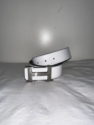 Vitt skärp med silverfärgat H-spänne - Snyggt vitt bälte med ett stilrent, silverfärgat H-format spänne. Bältet har en klassisk design och är tillverkat i skinn med svarta kanter och fina sömmar. Perfekt accessoar för att lyfta din outfit.