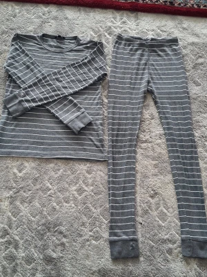Grå randig pyjamas i två delar - Mysig pyjamas i två delar med långärmad tröja och långa byxor. Setet är grått med tunna vita horisontella ränder och har en avslappnad passform. Perfekt för chillkvällar hemma. Materialet känns mjukt och stretchigt mot huden.