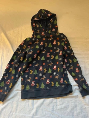 Julig hoodie med mönster från H&M för barn - Mörkblå hoodie från H&M med juligt all-over-mönster av tomtar, granar, snöflingor, renar, paket och texten 'HO HO HO'. Hoodien har långa ärmar och ribbade muddar. Perfekt för dig som vill maxa julkänslan med stil.