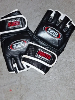 Svarta Fighter MMA-handskar i skinn - Svarta MMA-handskar från Fighter med vita detaljer och öppna fingrar. Tillverkade i skinn med kardborreband runt handleden för extra stöd. Perfekta för kampsport och träning. Snygg logga på ovansidan och robust design.